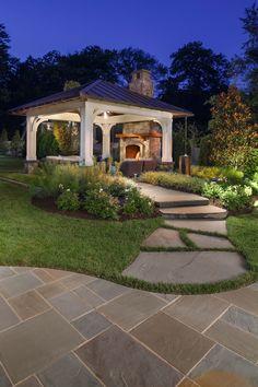 a gazebo in the middle of a garden at night with lights on and landscaping around it