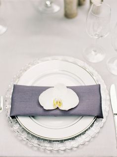 a white plate topped with a flower on top of a purple cloth covered place setting