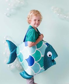 a little boy holding a large blue fish pillow in front of a wall with balloons