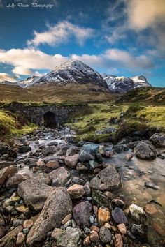 a stream running through a lush green field under a snow covered mountain range in the distance