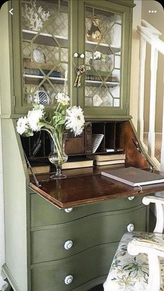 a desk with flowers in a vase on top of it next to a chair and bookshelf