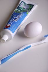 two toothbrushes sitting next to an egg on a white counter top, and one tube of toothpaste