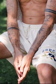 a man with tattoos sitting in the grass holding his hand up to his knee and looking down