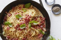 a pan filled with pasta and bacon on top of a white table next to two spoons