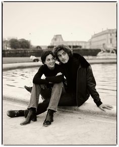 two people sitting next to each other on the ground near water and buildings in the background