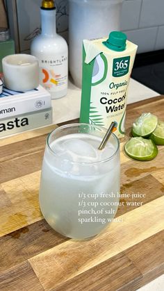 the ingredients to make an alcoholic cocktail are displayed on a cutting board, including limes and a carton of coconut water