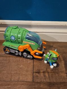 a green toy truck with an orange cat next to it on a wooden floor in front of a blue wall