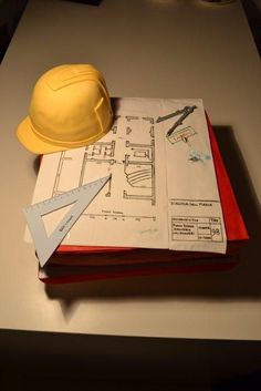 a yellow hard hat sitting on top of an open book next to construction drawings and scissors