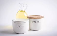 two white containers with labels on them next to a glass jug and a wooden lid