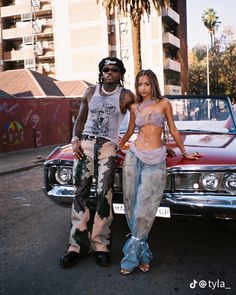 two people standing in front of a car on the street with palm trees behind them