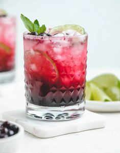 a close up of a drink in a glass on a table with limes and blueberries