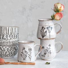 three ceramic mugs with designs on them next to a wooden spoon and orange flower