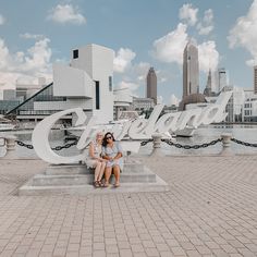 two women are sitting on a bench in front of the cleveland sign, with city buildings in the background