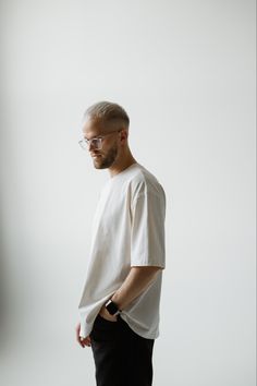 a man with glasses standing in front of a white wall wearing black pants and a white t - shirt