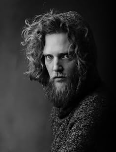 a black and white photo of a man with long curly hair wearing a coat looking at the camera