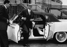 two men in suits standing next to a car with its doors open and the door opened