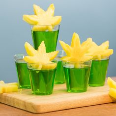 four shot glasses filled with green liquid and cut up pineapples on a cutting board