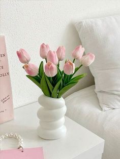 a white vase filled with pink tulips sitting on top of a table next to a book