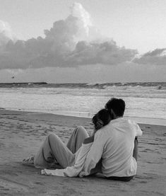 two people sitting on the beach looking out at the water and clouds in the sky