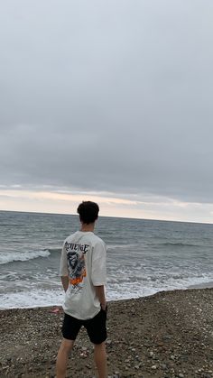 a young man standing on top of a beach next to the ocean under a cloudy sky