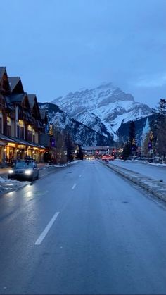 cars are driving down the street in front of some buildings with snow covered mountains behind them