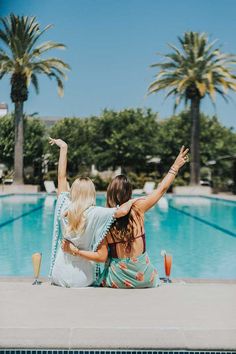 two women sitting on the edge of a swimming pool with their arms in the air