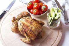 a wooden plate topped with a chicken next to a bowl of cucumbers and tomatoes