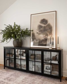 a black cabinet with glass doors and vases on top in front of a painting