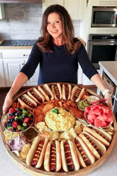 a woman standing in front of a large platter of hotdogs and fruit