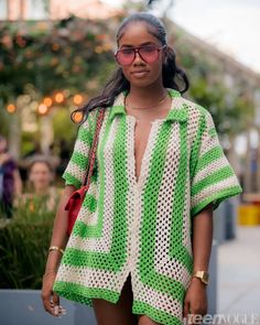 a woman in green and white crocheted shirt walking down the street with her hand on her hip