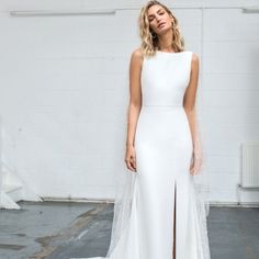 a woman standing in front of a white wall wearing a wedding dress with a high slit