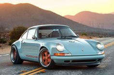 an old blue porsche sits on the side of the road in front of some mountains