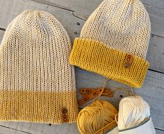 two knitted hats sitting on top of a wooden floor next to balls of yarn