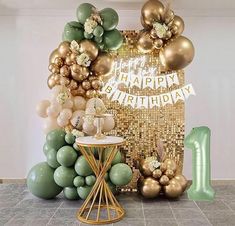 a table topped with balloons and a number one sign next to a wall covered in gold, green and white balloons