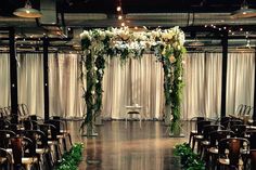 an indoor wedding ceremony setup with white flowers and greenery on the back drop cloth drapes