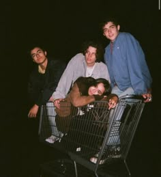 three young men are sitting on top of a shopping cart in the dark, one is leaning against the fence