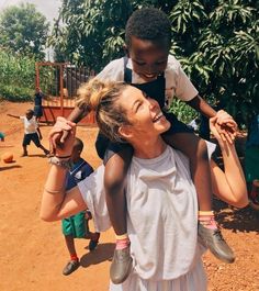 a woman is carrying a child on her shoulders while others watch from the dirt ground