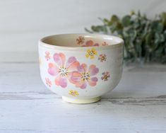 a white bowl with pink and yellow flowers painted on the side sitting next to a green plant