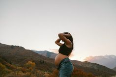 a pregnant woman standing in the mountains with her hands on her head looking at the sky