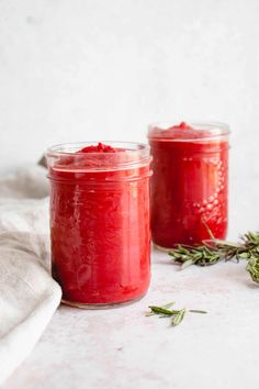 two jars filled with red food sitting on top of a white tablecloth next to a sprig of rosemary