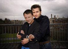 two men standing next to each other in front of a metal fence with buildings behind them