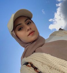 a woman wearing a hat and scarf standing in front of a blue sky with clouds