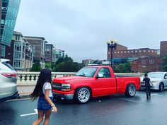 a woman walking across a parking lot next to a red pick up truck and other cars