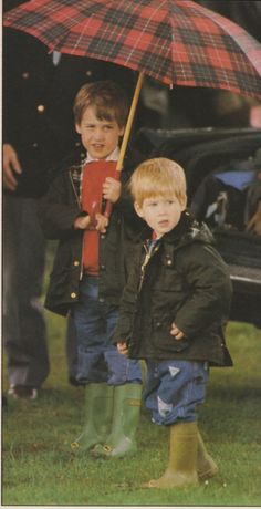 two young boys standing under an umbrella in the grass with other people behind them and one boy wearing rubber boots