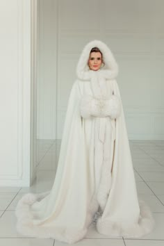 a woman in a white gown and fur coat standing on the floor with her arms crossed