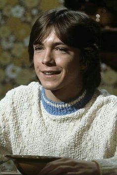 a smiling woman sitting at a table with a plate in front of her, wearing a white sweater