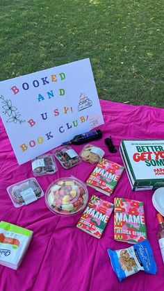 a table topped with books and snacks on top of a pink cloth covered picnic blanket