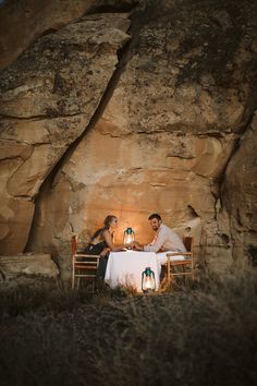 a man and woman sitting at a table in front of a large rock formation with candles lit