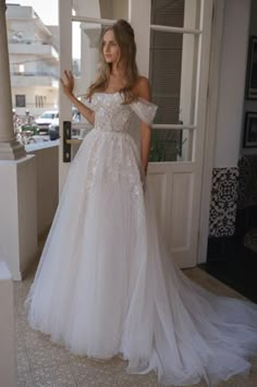 a woman standing in front of a door wearing a wedding dress with an off the shoulder design