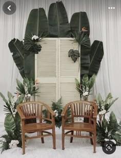 two wooden chairs sitting next to each other in front of a window with large green leaves